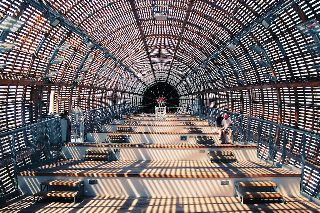 Tunnel-like structure with latticed metal framework creating striped light patterns on floor.