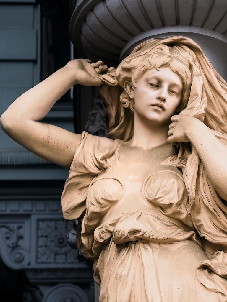 Marble statue of a woman with flowing hair and draped clothing, hand raised to head.